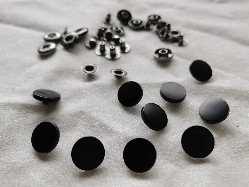 Puede incluir: Una colecci&oacute;n de botones a presi&oacute;n de metal negro, algunos con detalles plateados, sobre un fondo de tela blanca.