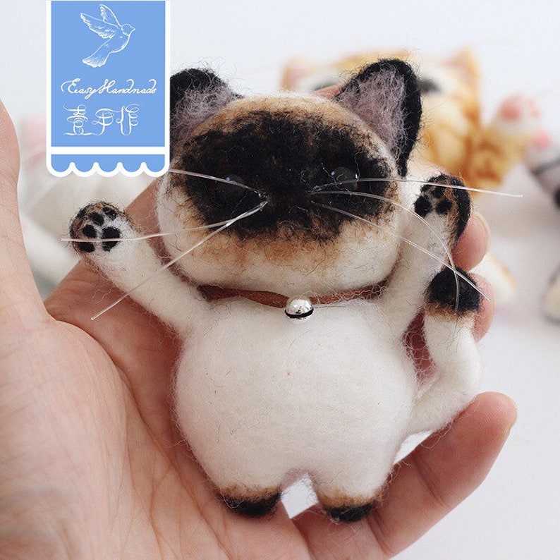 May include: A white, brown, and black needle felted cat with a silver bell on a brown collar. The cat is standing on its hind legs with its front paws raised. The cat is being held in a person's hand.