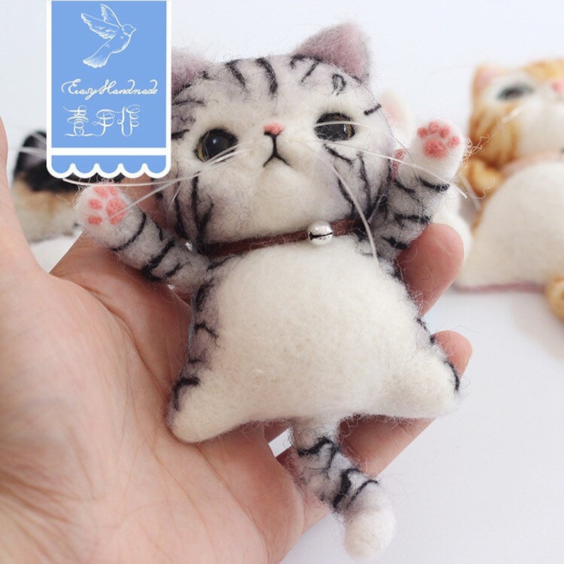 May include: A small, gray and white felted cat with black stripes, a brown collar with a silver bell, and pink paw pads. The cat is sitting up with its paws raised and its tail curled behind it.
