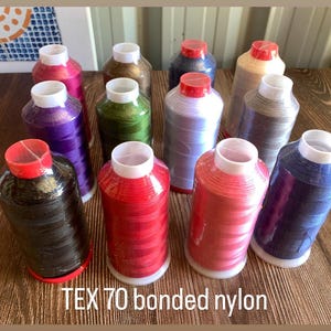 May include: Assortment of spools of thread in various colours, including red, green, blue, purple, brown, and beige. The spools are labelled "TEX 70 bonded nylon".