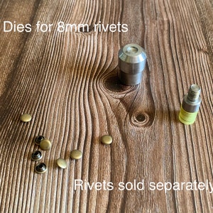 Op de afbeelding: Twee metalen matrijzen voor het zetten van 8 mm klinknagels, samen met een selectie van acht messing klinknagels.  "Dies for 8mm rivets" en "Rivets sold separately" staan op het houten oppervlak.