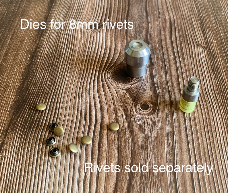 Op de afbeelding: Twee metalen matrijzen voor het zetten van 8 mm klinknagels, samen met een selectie van acht messing klinknagels.  "Dies for 8mm rivets" en "Rivets sold separately" staan op het houten oppervlak.