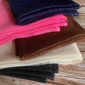May include: Five pieces of mesh fabric in different colours, including pink, brown, white, black, and navy blue. The fabric is folded and stacked on a wooden surface.