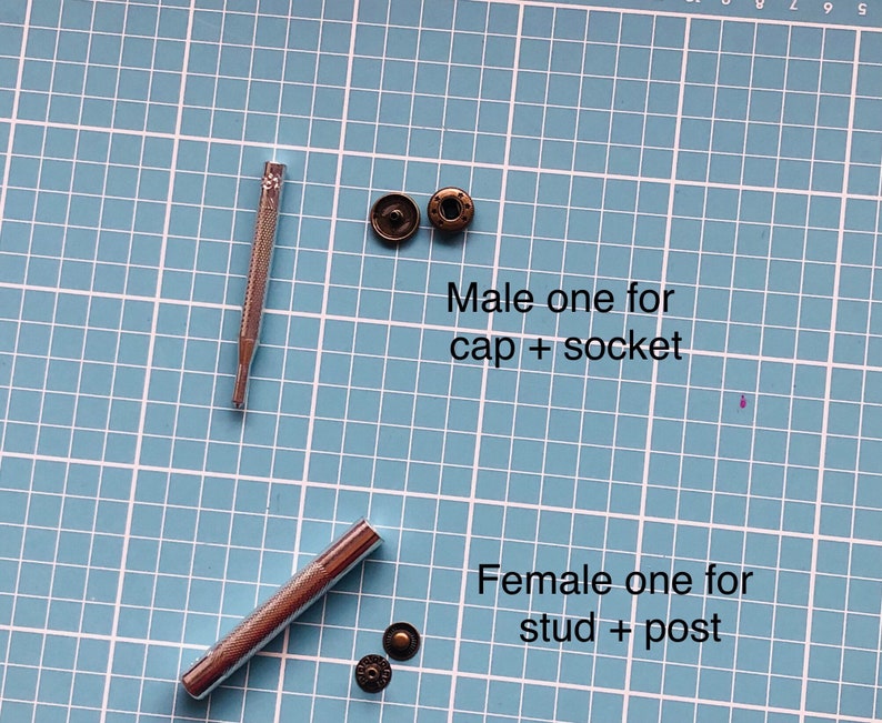 Puede incluir: Dos herramientas de metal para colocar broches a presi&oacute;n, una para broches a presi&oacute;n masculinos y otra para broches a presi&oacute;n femeninos. Las herramientas est&aacute;n sobre una estera de corte azul. Tambi&eacute;n hay cuatro broches a presi&oacute;n de metal, dos masculinos y dos femeninos.