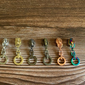 Könnte beinhalten: Sechs Metall-Reißverschlussanhänger in verschiedenen Ausführungen: Silber, Gold, Bronze, Kupfer und schillernd. Jeder Anhänger hat einen Ring am unteren Ende. Auf einer Holzoberfläche angeordnet.