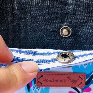 Puede incluir: Primer plano de una tela a rayas blancas y azules con un bot&oacute;n marr&oacute;n. La tela est&aacute; unida a una tela blanca con un estampado floral. Una etiqueta de cuero marr&oacute;n con la palabra "Handmade" est&aacute; unida a la tela.