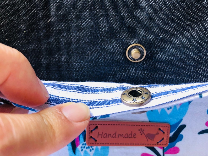 Puede incluir: Primer plano de una tela a rayas blancas y azules con un bot&oacute;n marr&oacute;n. La tela est&aacute; unida a una tela blanca con un estampado floral. Una etiqueta de cuero marr&oacute;n con la palabra "Handmade" est&aacute; unida a la tela.