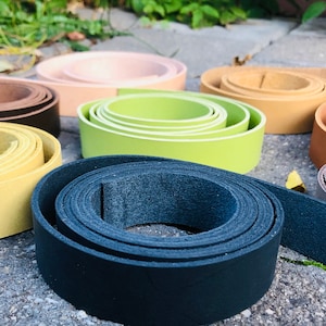 May include: Assortment of leather straps in various colours, including black, brown, beige, yellow, green, and grey. The straps are rolled up and arranged on a grey concrete surface.