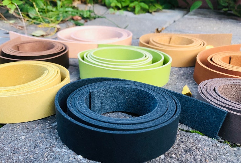 K&ouml;nnte beinhalten: Sortiment aus Lederriemen in verschiedenen Farben, darunter Schwarz, Braun, Beige, Gelb, Gr&uuml;n und Grau. Die Riemen sind aufgerollt und auf einer grauen Betonfl&auml;che angeordnet.