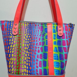 K&ouml;nnte beinhalten: Eine leuchtend bunte Tragetasche mit einem regenbogenfarbenen Krokodilledermuster. Die Tasche hat einen Rei&szlig;verschluss und zwei Ledergriffe mit silbernen Nieten. Die Tasche ist mit einem leuchtend pinkfarbenen Stoff gef&uuml;ttert.