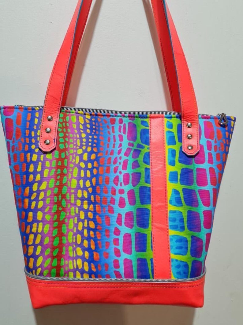 K&ouml;nnte beinhalten: Eine leuchtend bunte Tragetasche mit einem regenbogenfarbenen Krokodilledermuster. Die Tasche hat einen Rei&szlig;verschluss und zwei Ledergriffe mit silbernen Nieten. Die Tasche ist mit einem leuchtend pinkfarbenen Stoff gef&uuml;ttert.