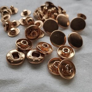 Puede incluir: Una colecci&oacute;n de botones a presi&oacute;n de metal dorado, incluyendo las piezas superior e inferior. Los botones a presi&oacute;n est&aacute;n dispuestos en un patr&oacute;n aleatorio sobre una superficie blanca.