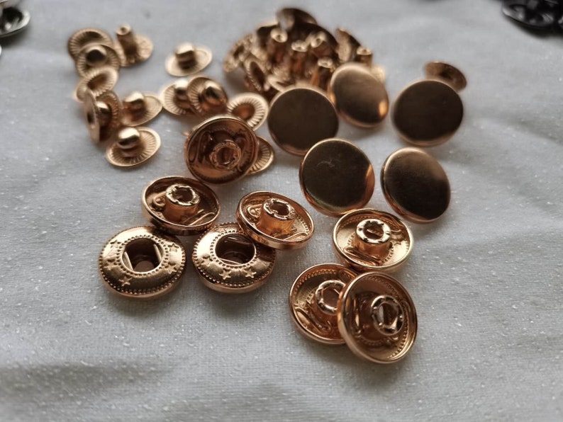 Puede incluir: Una colecci&oacute;n de botones a presi&oacute;n de metal dorado, incluyendo las piezas superior e inferior. Los botones a presi&oacute;n est&aacute;n dispuestos en un patr&oacute;n aleatorio sobre una superficie blanca.