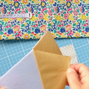 Puede incluir: Primer plano de una persona sosteniendo un trozo de papel marr&oacute;n con un trozo de papel blanco debajo. El papel est&aacute; sobre una estera de corte azul con una tela floral blanca y rosa en el fondo.
