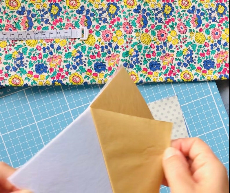 Puede incluir: Primer plano de una persona sosteniendo un trozo de papel marr&oacute;n con un trozo de papel blanco debajo. El papel est&aacute; sobre una estera de corte azul con una tela floral blanca y rosa en el fondo.