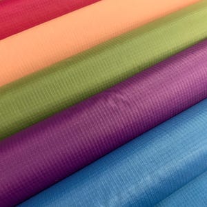May include: Multiple rolls of ripstop fabric arranged diagonally, featuring a grid pattern. The colours include vibrant pink, soft peach, olive green, deep purple, and bright blue. Each roll displays a distinct texture with a visible square weave.