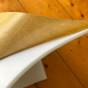 Puede incluir: Una l&aacute;mina de espuma blanca con un respaldo de papel marr&oacute;n. El papel est&aacute; parcialmente despegado para revelar la espuma.