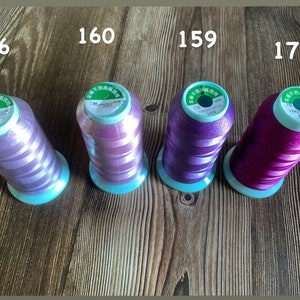 May include: Four spools of embroidery thread in different shades of purple and pink. The spools are labeled with numbers 156, 160, 159, and 176.