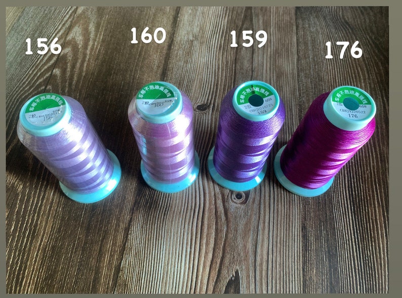 May include: Four spools of embroidery thread in different shades of purple and pink. The spools are labeled with numbers 156, 160, 159, and 176.