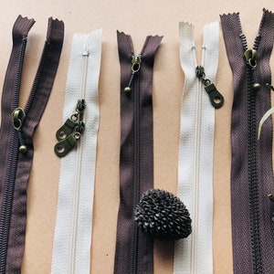 May include: Five different zipper closures in various colors and styles. The zippers are laid out horizontally on a brown surface. The zippers are all closed and have metal pulls.