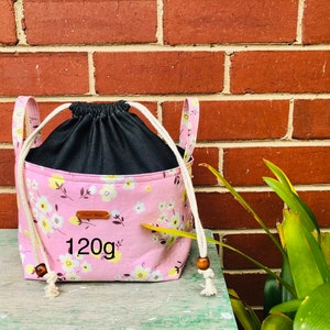 K&ouml;nnte beinhalten: Rosaer Kordelzugbeutel mit Blumendruck und schwarzem Futter. Die Tasche hat ein braunes Leder-Etikett mit der Aufschrift "Hand Made" und "120g".