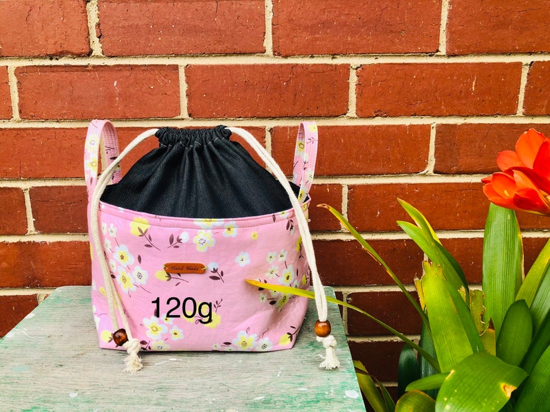 K&ouml;nnte beinhalten: Rosaer Kordelzugbeutel mit Blumendruck und schwarzem Futter. Die Tasche hat ein braunes Leder-Etikett mit der Aufschrift "Hand Made" und "120g".