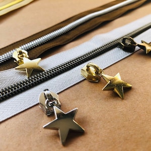May include: Four different zipper pulls with star-shaped charms. The charms are gold and silver metal. The zippers are brown, white, and silver.