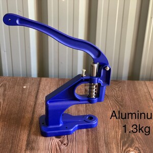 Op de afbeelding: Een blauwe metalen knopenpers met een hendel. De machine is gemaakt van aluminium en weegt 1,3 kg.