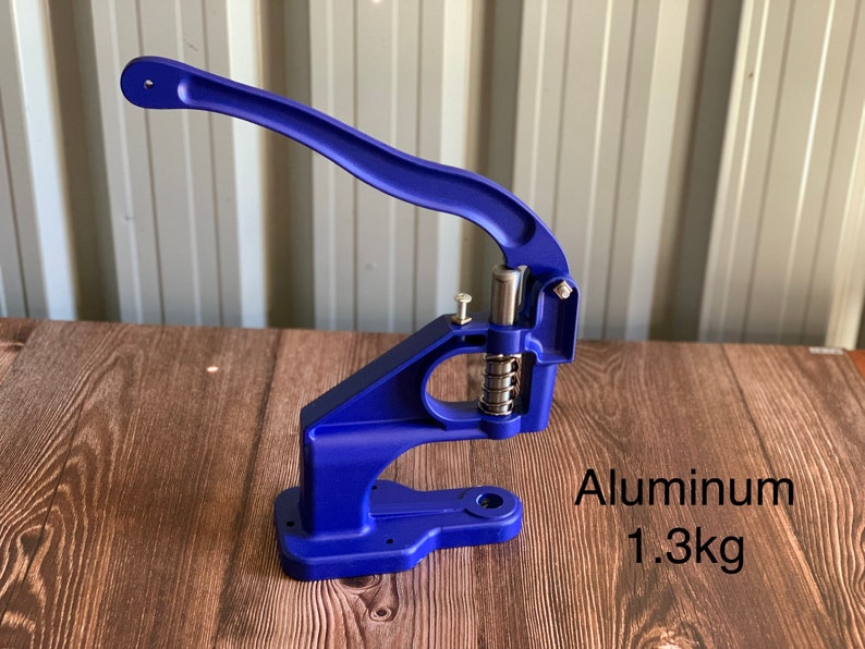 Op de afbeelding: Een blauwe metalen knopenpers met een hendel. De machine is gemaakt van aluminium en weegt 1,3 kg.