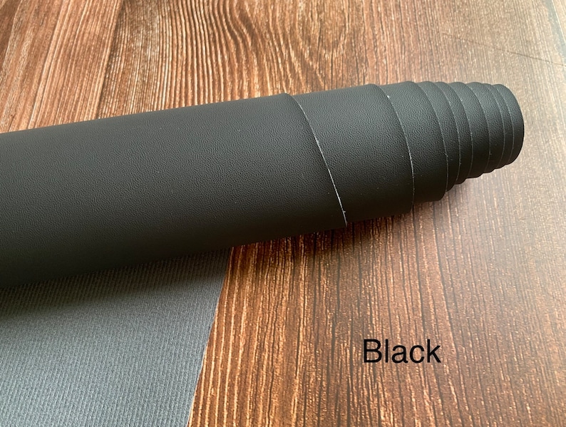 Puede incluir: Una l&aacute;mina de cuero sint&eacute;tico negro enrollada. El cuero tiene una superficie texturizada. El texto "Black" est&aacute; escrito en una superficie de madera debajo del cuero.