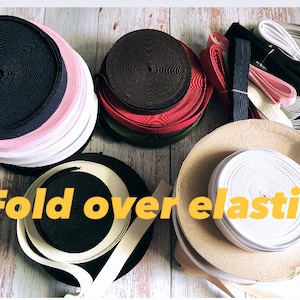 Peut inclure: Assortiment d'élastique à ourlet en différentes couleurs et largeurs. L'élastique est enroulé et empilé sur une surface en bois. Le texte "Fold over elastic" est écrit en lettres jaunes sur l'image.