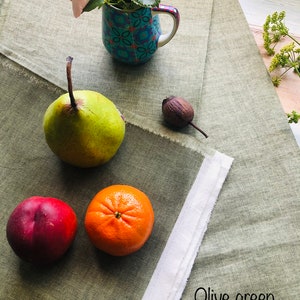 K&ouml;nnte beinhalten: Eine Stillleben mit einer rosa Rose in einer Tasse mit blau-wei&szlig;em Muster, einer gr&uuml;nen Birne, einem roten Pfirsich und einer Orange auf einer gr&uuml;nen Tischdecke aus Leinen. Der Text "Olive green" ist auf der Tischdecke geschrieben.