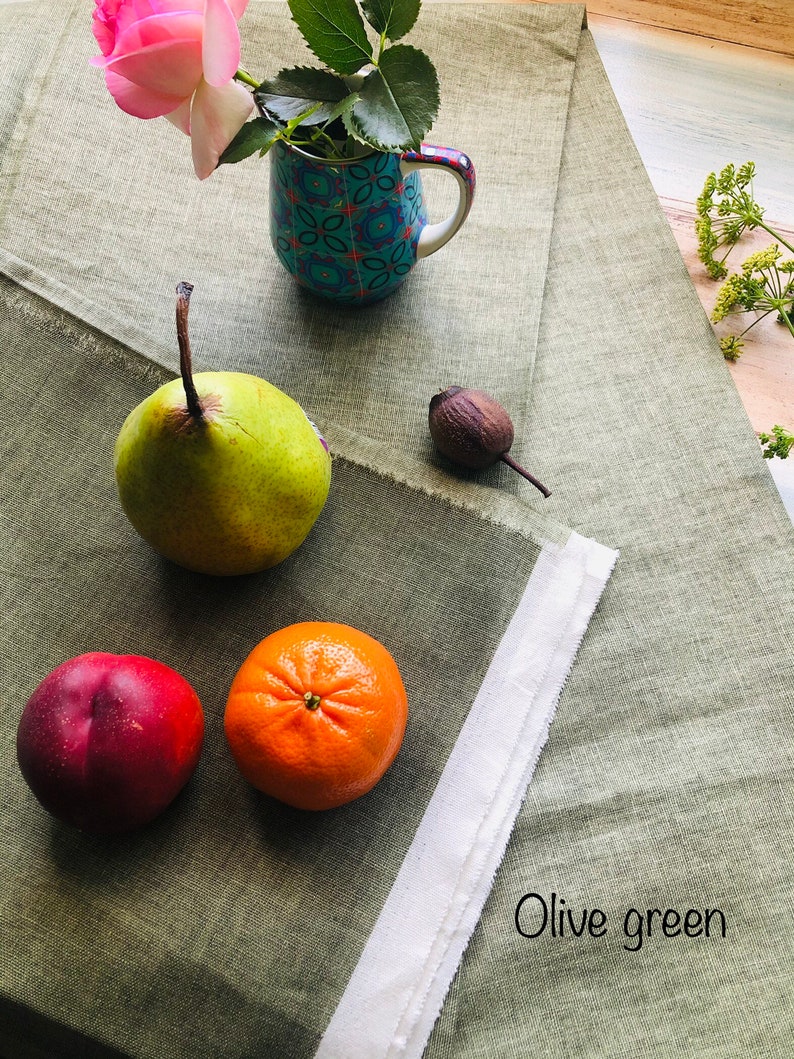 K&ouml;nnte beinhalten: Eine Stillleben mit einer rosa Rose in einer Tasse mit blau-wei&szlig;em Muster, einer gr&uuml;nen Birne, einem roten Pfirsich und einer Orange auf einer gr&uuml;nen Tischdecke aus Leinen. Der Text "Olive green" ist auf der Tischdecke geschrieben.
