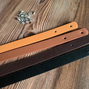 Peut inclure: Quatre lanières en cuir de différentes couleurs : marron, beige, marron foncé et noir. Les lanières sont disposées sur une surface en bois.