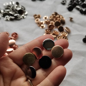 May include: Assortment of round metal snaps in various colors, including gold, silver, black, and copper. The snaps are displayed on a hand and on a white surface.