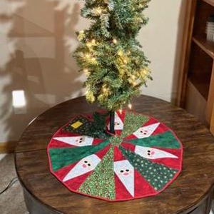 May include: A small, artificial Christmas tree with warm white lights stands on a round, wooden table. A colorful tree skirt with Santa Claus faces and red and green sections surrounds the base of the tree. A framed picture hangs on the wall.
