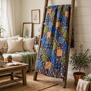 May include: A patchwork quilt featuring a repeating pattern of square fabric blocks in shades of blue, green, orange, and a floral print. The quilt is draped over a tall, rustic wooden ladder, showcasing its quilted texture and stitched grid design.