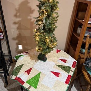 May include: A small, artificial Christmas tree with warm white lights stands on a round table. The tree is surrounded by a quilted tree skirt with a pattern of green Christmas trees, red gnome hats, and white squares.