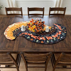 May include: A curved Halloween-themed table runner featuring a pattern of pumpkins, ghosts, and spiderwebs in orange, black, and white. A glass bowl filled with assorted candies sits in the center, flanked by two small white ghost figurines.