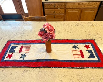 May include: A patriotic table runner with a white center, red and blue borders, and star and flag appliqués. A brown vase holds red, white, and blue flowers. The runner is approximately 12 inches wide and 36 inches long.