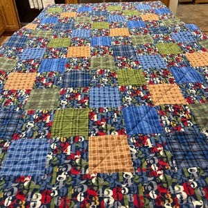 May include: A patchwork quilt with a variety of fabrics in blue, green, brown, and red. The quilt features squares of plaid, solid colors, and a fabric with white numbers. The quilt is a handmade item.