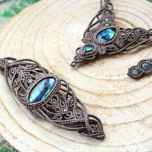 Può includere: Un set di gioielli in macramè realizzato con cordino marrone intrecciato in motivi complessi con pietre di labradorite blu iridescente incastonate. Il set include una collana, un bracciale e un piccolo accessorio, tutti con dettagliati nodi.