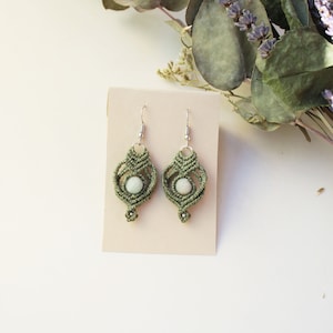 Può includere: Un paio di orecchini in macramè verde oliva con una perla rotonda verde chiaro al centro. Gli orecchini presentano intricati motivi a cordoncino annodato e sono appesi a ganci in metallo color argento su un cartoncino rettangolare beige.