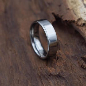 Könnte beinhalten: Ein polierter, silberfarbener Ring mit flacher Oberseite und abgerundeten Kanten. Der Ring liegt auf einer strukturierten, dunkelbraunen Holzoberfläche. Die glatte Oberfläche des Rings reflektiert das Licht.