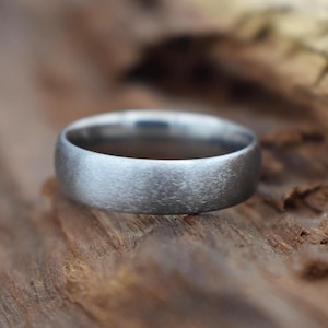 May include: A close-up of a brushed silver ring with a rounded band. The ring is set against a textured, wooden surface. The ring's surface has a matte finish, reflecting soft light.