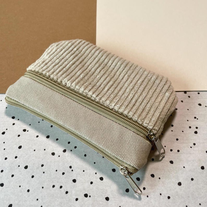 May include: A beige corduroy pencil case with a canvas base and a zipper closure. The case has a rectangular shape and a soft, textured appearance. The zipper is silver-toned.