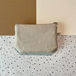 May include: A beige canvas zippered pouch, rectangular in shape. The pouch has a silver-colored zipper and a small loop on the side. The background is a mix of brown, cream, and a speckled black and white pattern.