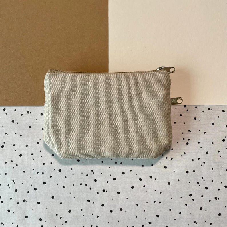 May include: A beige canvas zippered pouch, rectangular in shape. The pouch has a silver-colored zipper and a small loop on the side. The background is a mix of brown, cream, and a speckled black and white pattern.