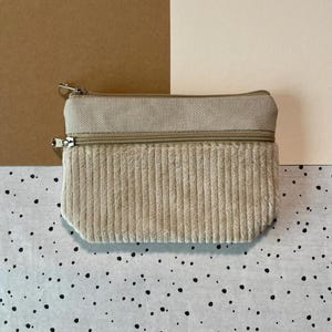 May include: A beige corduroy and canvas zippered pouch. The top section is canvas, and the bottom is corduroy. The pouch has two separate zippered compartments with silver-colored metal zippers and pulls. The background is a mix of neutral colors.