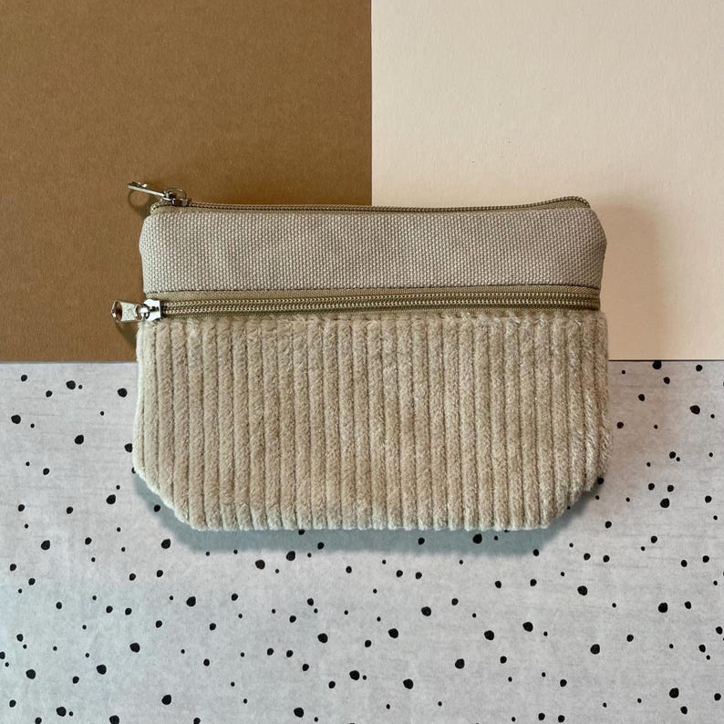May include: A beige corduroy and canvas zippered pouch. The top section is canvas, and the bottom is corduroy. The pouch has two separate zippered compartments with silver-colored metal zippers and pulls. The background is a mix of neutral colors.